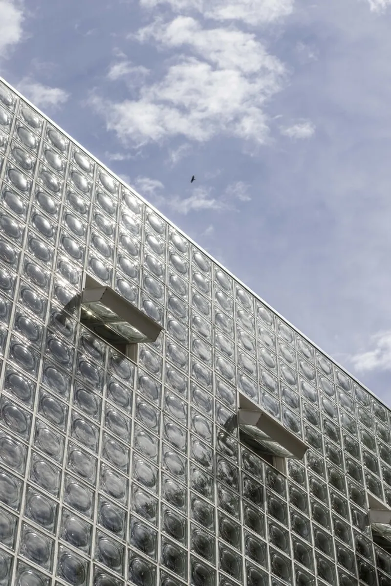 Glass block facade with rectangular window openings and a bird flying overhead in blue sky