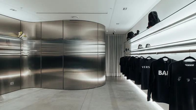 Cylindrical stainless steel volume with horizontal lit recesses adjacent to white shelving displaying folded apparel