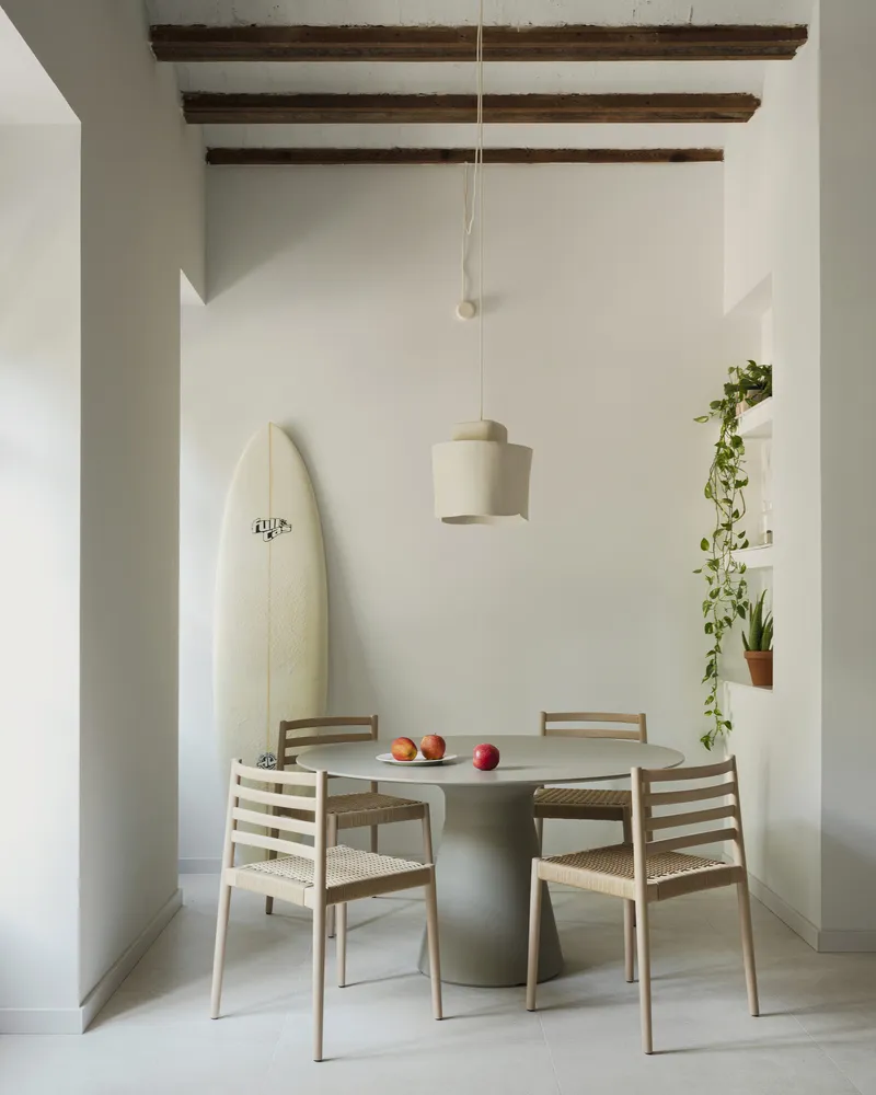 Dining area with round pedestal table and surfboard leaning against white wall beneath timber beams