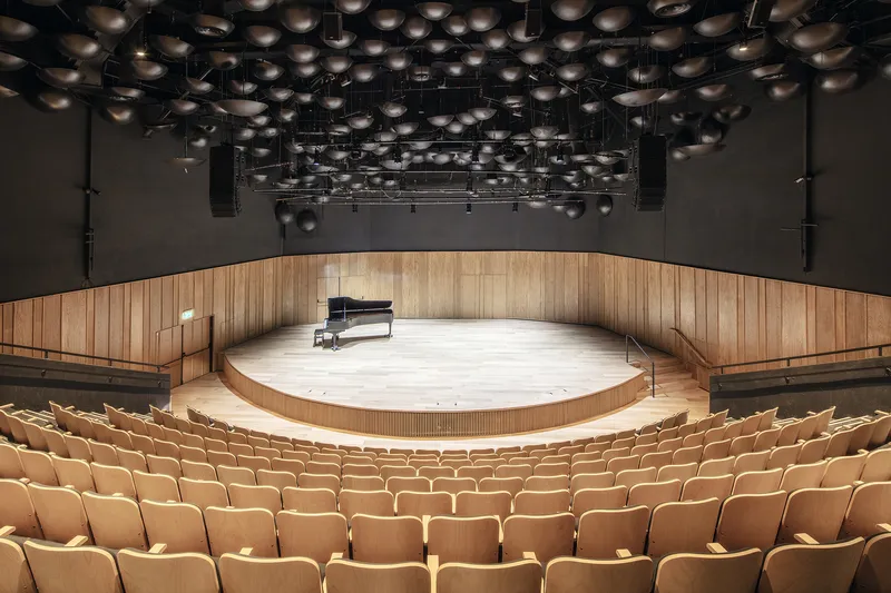Concert hall with circular timber stage, rows of seating, and overhead array of acoustic reflectors