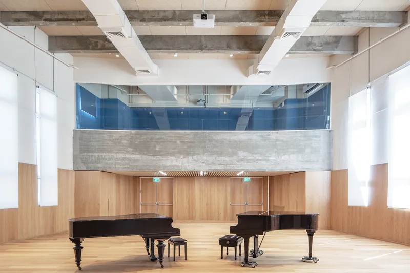 Rehearsal room with two grand pianos, exposed concrete beams, and upper-level observation gallery with blue glass