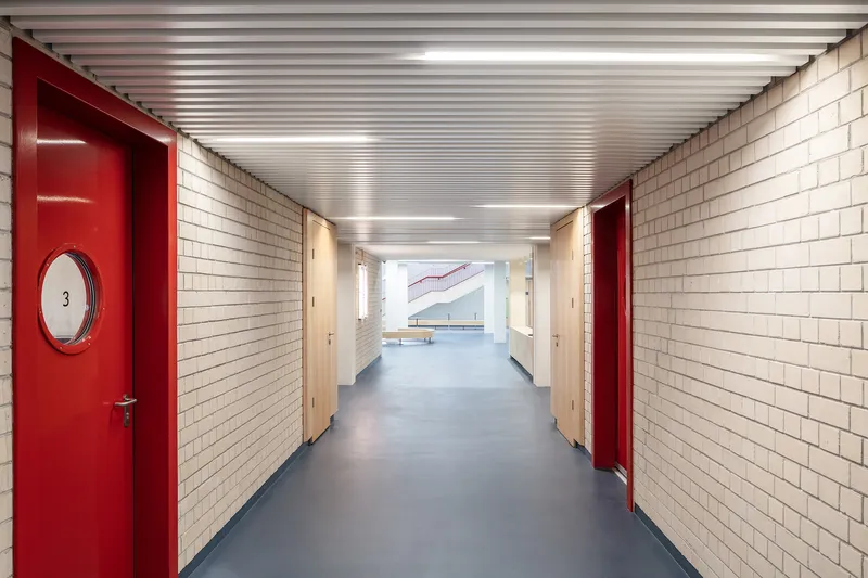 Corridor with white tile walls, red doors with circular windows, and linear ceiling lighting