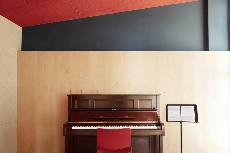 Corner detail showing upright piano against plywood wall beneath red acoustic ceiling panels and grey soffit