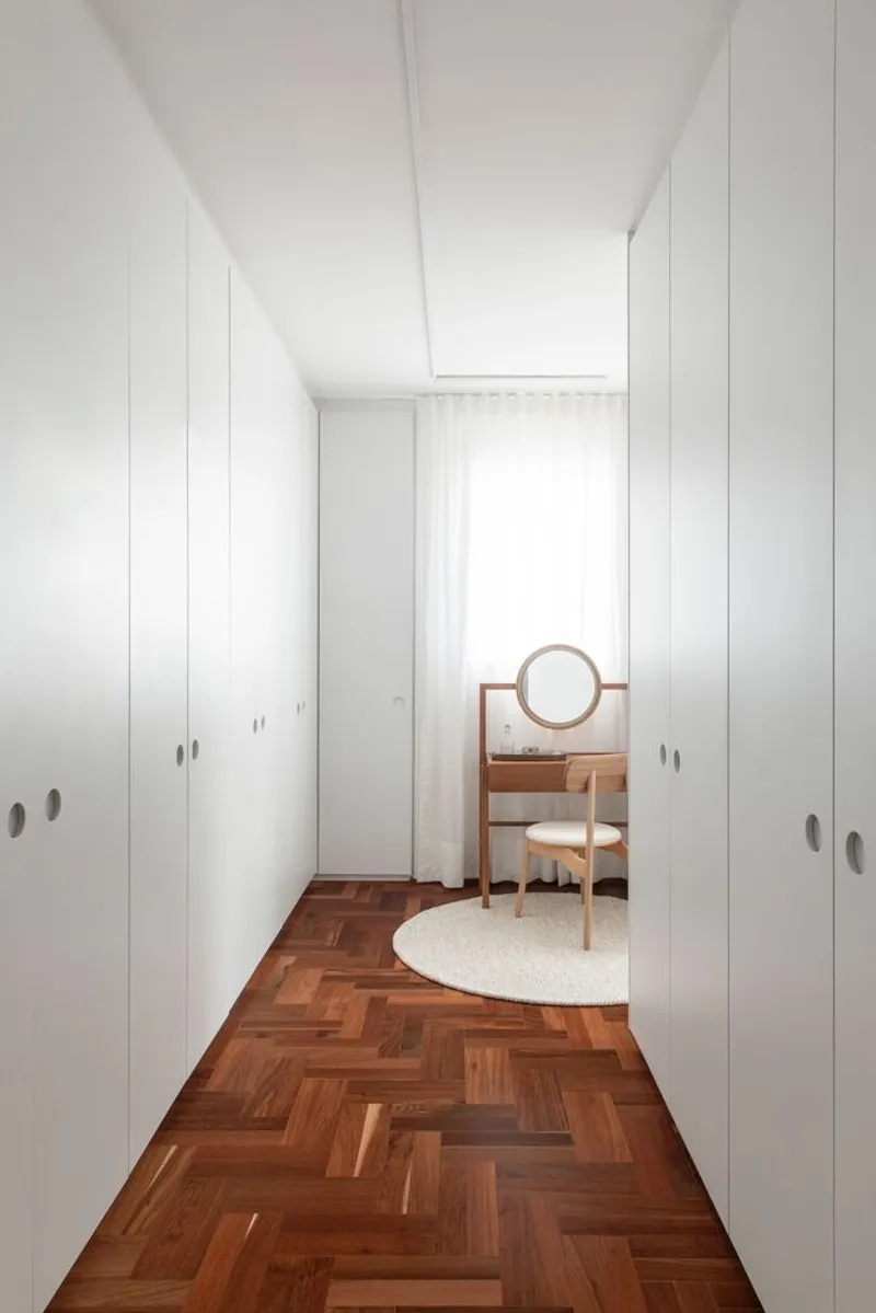 Narrow corridor with white storage walls flanking herringbone parquet and vanity visible through doorway