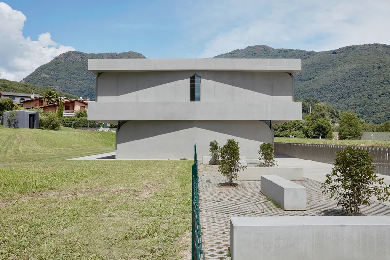 Facade with stacked curved voids and entrance courtyard with planted beds and checkerboard paving under summer clouds