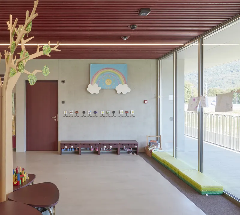 Classroom with slatted maroon ceiling, glazed wall opening to terrace, and wall-mounted rainbow artwork above storage cubbies