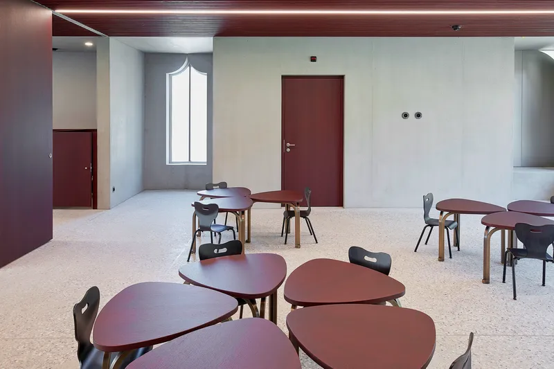 Room with terrazzo flooring and organic-shaped tables scattered among maroon doors and a tall arched window