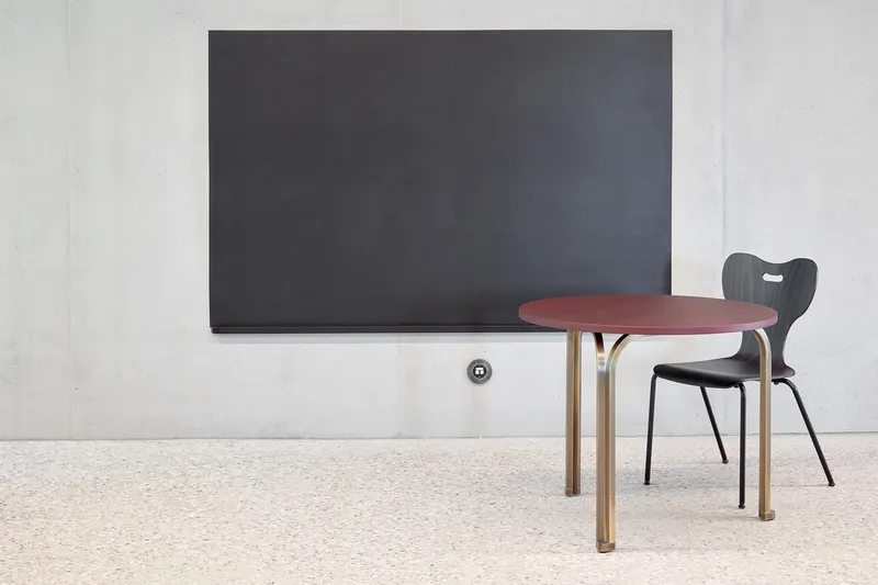 Detail of a curved table with tubular legs and a plywood chair against a blackboard on concrete wall