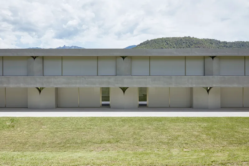 Two-story concrete facade with recessed openings and arched spandrels under a partly cloudy sky and hills