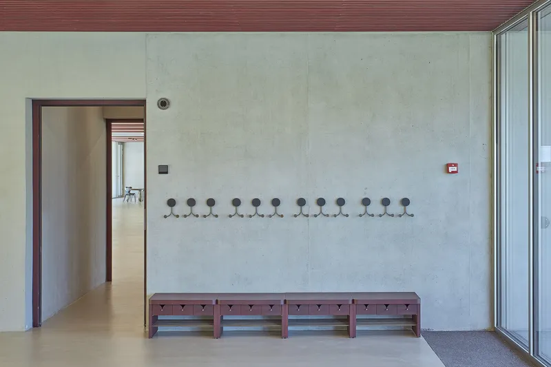 Entry corridor with row of coat hooks above burgundy benches along a pale concrete wall