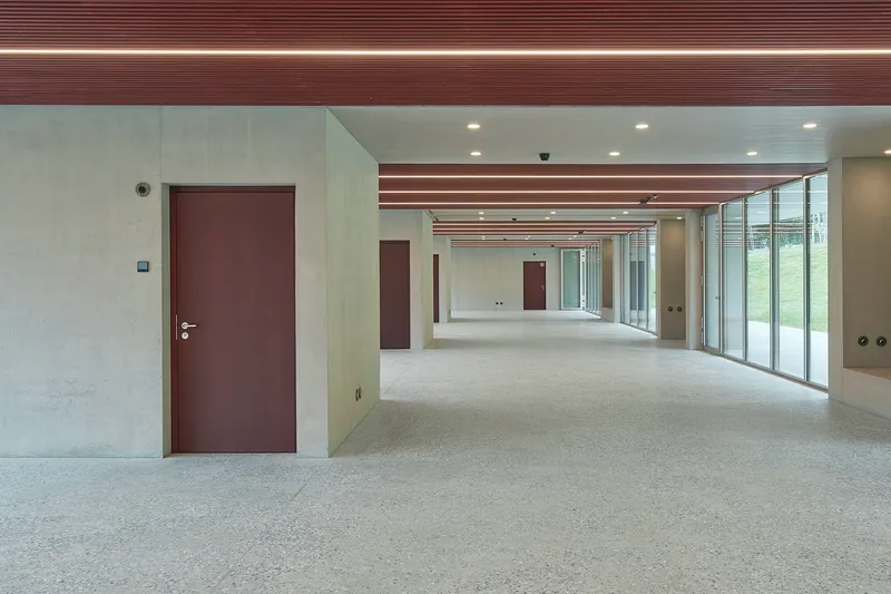 Long corridor with terrazzo floor, exposed maroon ceiling beams and glazed wall overlooking lawn
