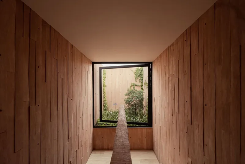 Narrow hallway with timber walls converging toward a glazed opening framing a narrow planted garden