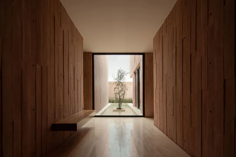Timber-lined corridor leading to a glazed opening framing a small tree in an interior courtyard
