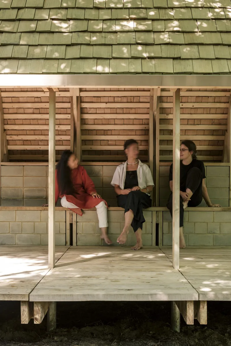 Covered porch with timber columns and brick infill walls where three people sit in dappled sunlight