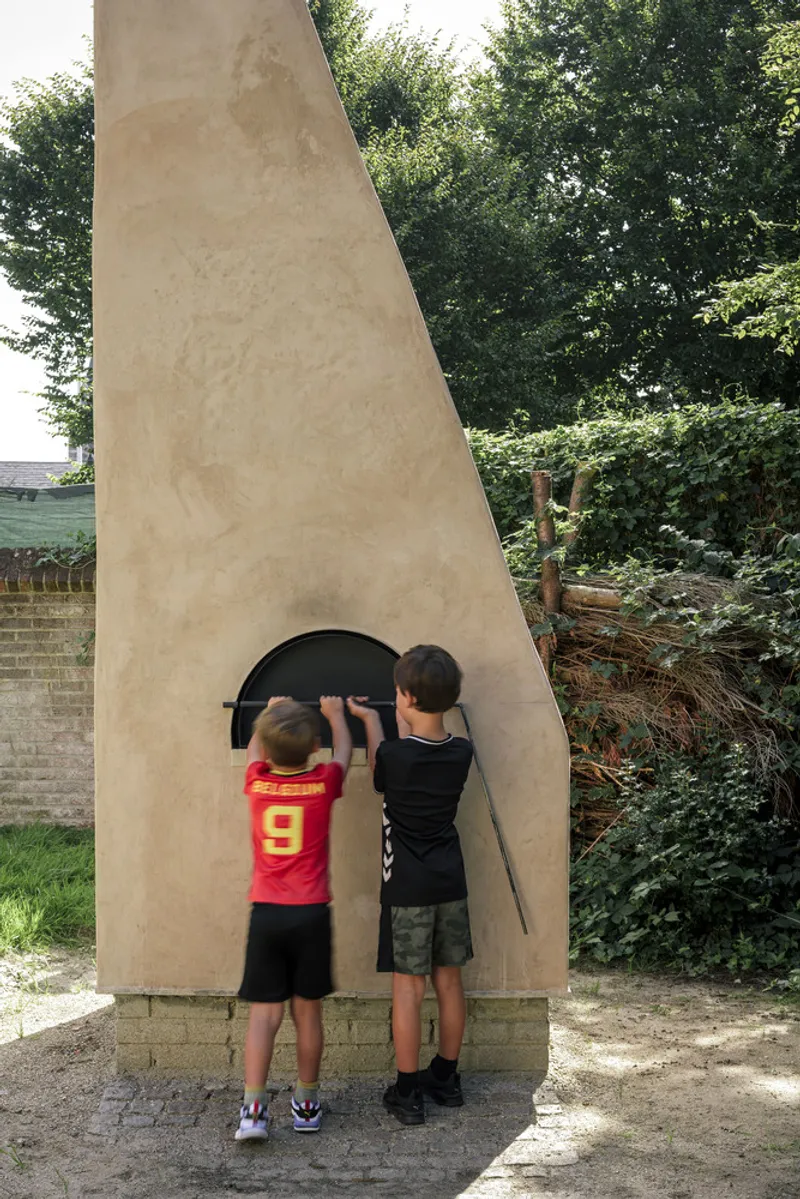 Tapered outdoor oven with arched opening on brick base as two children peer inside the firebox