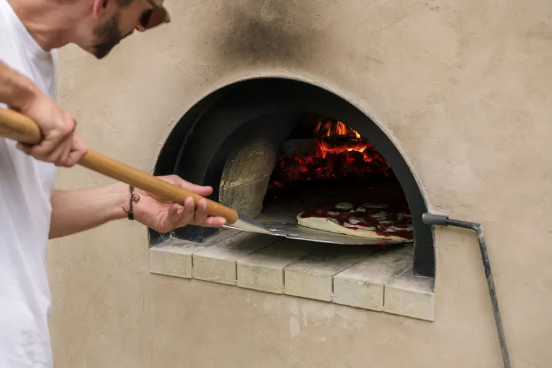 Arched oven opening with glowing embers as hands slide a pizza peel into the firebox