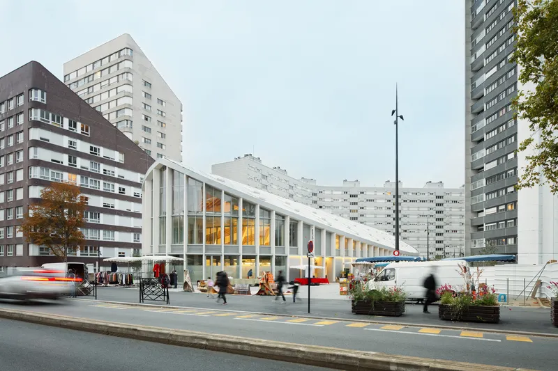 Street view of the glazed pavilion with vertical louvers nestled among residential towers with pedestrians passing
