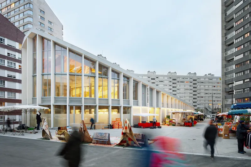 Glazed facade with arched openings and interior lighting at dusk with pedestrians and market stalls