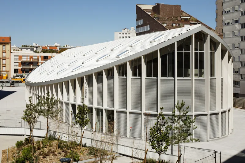 Oval volume with white vertical louvers and barrel vault roof surrounded by residential towers and young trees