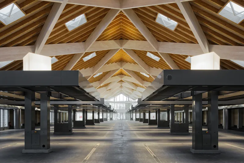 Empty interior showing black steel columns beneath layered timber trusses with triangular roof vents