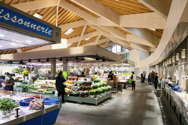 Interior market hall with exposed timber trusses and white beams above produce stalls and shoppers