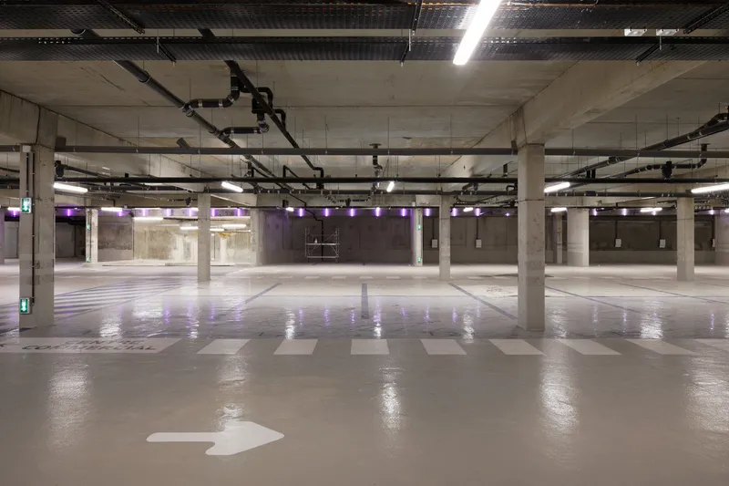 Underground parking level with concrete columns, exposed mechanical piping, and painted wayfinding arrows on the floor
