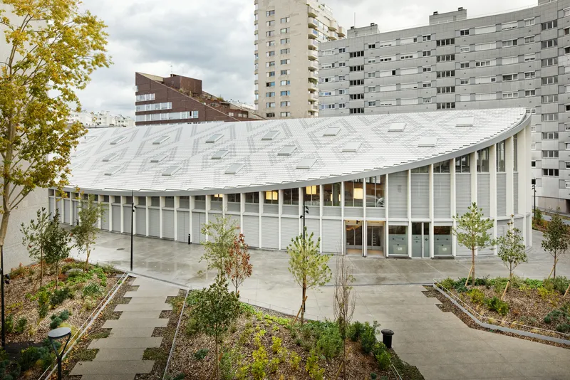 Curved white roof with skylights rising above a plaza with newly planted trees and residential towers beyond