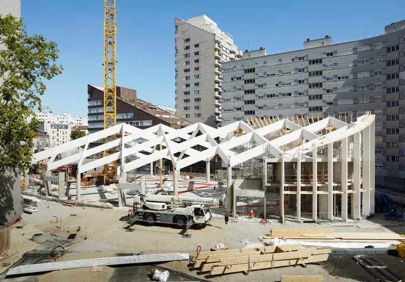 Construction site view of the white zigzag roof structure supported by columns, with residential towers in the background