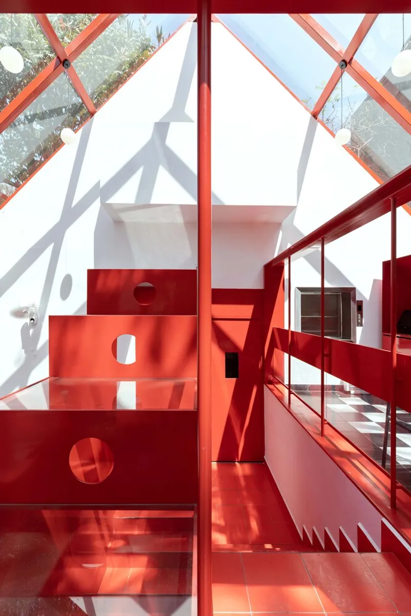 Red steel staircase with perforated treads rising through a double-height space with glazed diagonal bracing