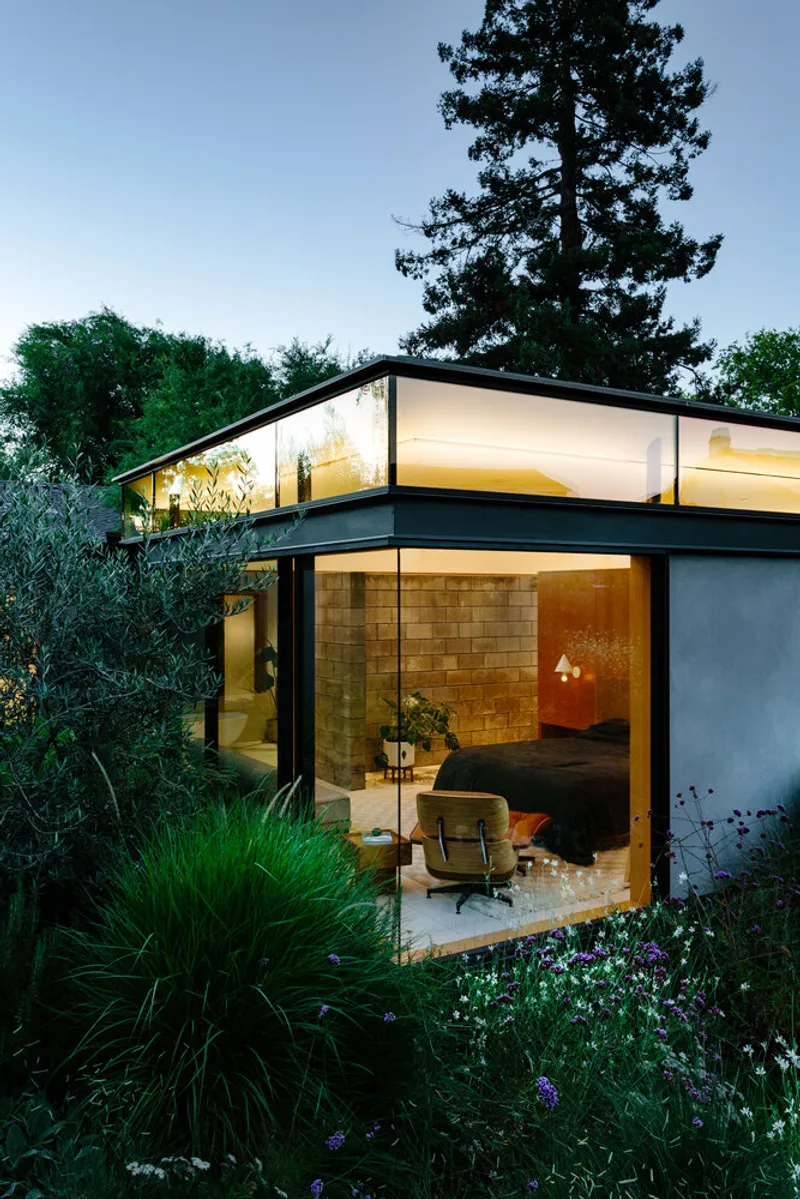Glass-walled living room with translucent clerestory band illuminated at dusk among grasses and trees