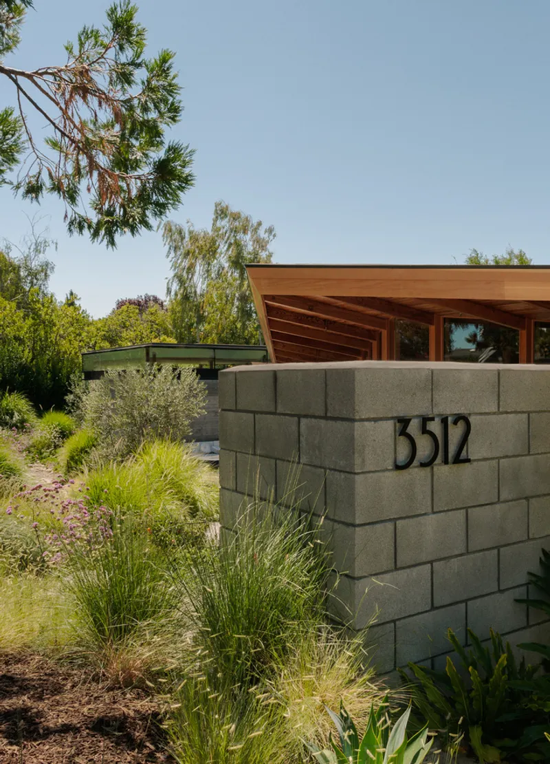 Concrete block entry wall with house numbers beside planted grasses under a timber canopy