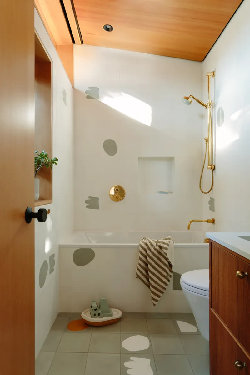 Bathroom with white tiled tub surround, brass fixtures and plywood ceiling lit by morning sunlight