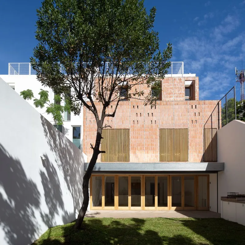 Rear facade of terracotta brick with vertical timber shutters and glazed pavilion below a mature tree