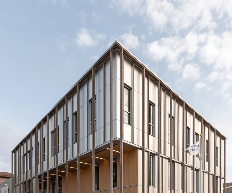 Corner elevation showing the two-story timber louvered facade with recessed ground-floor glazing beneath scattered clouds