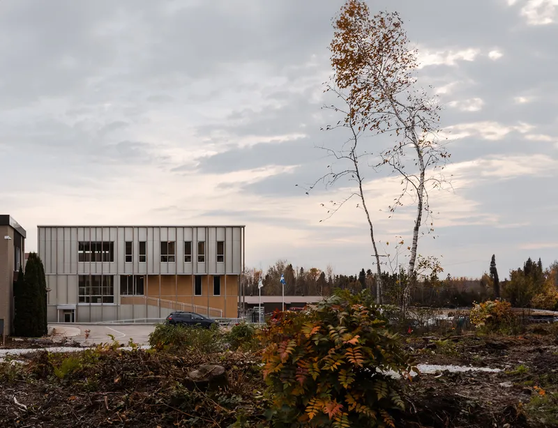 Two-storey building with vertical metal cladding seen across a cleared lot with autumn foliage and birches