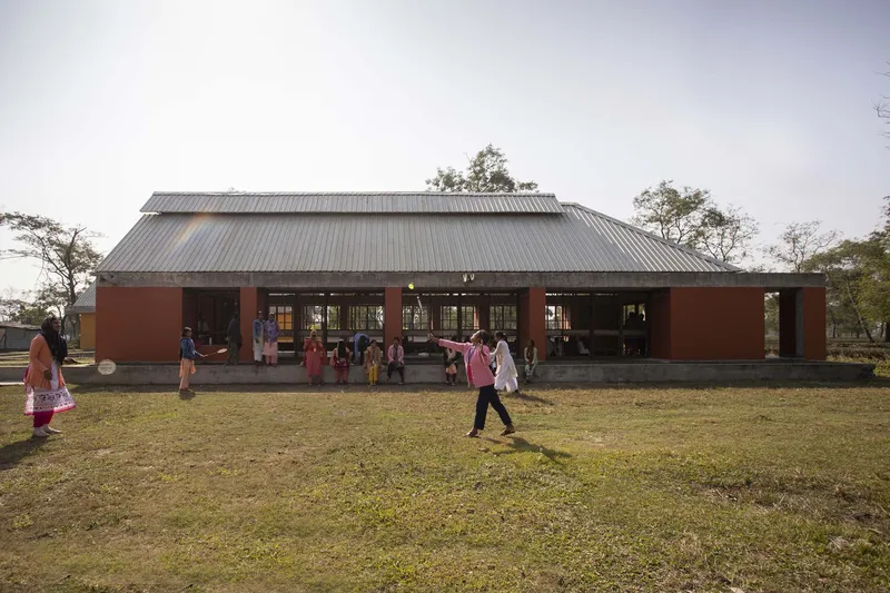 Red masonry pavilion with corrugated metal roof and open colonnade facing a lawn with children playing