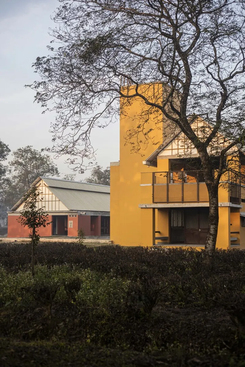 Yellow two-story block beside a low red pavilion framed by bare tree branches at dusk