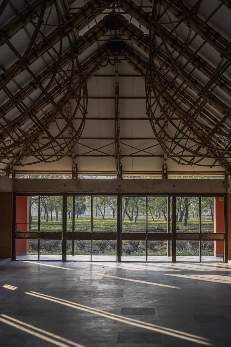 Interior view of exposed timber truss structure over a dark floor looking toward full-height glazing and trees