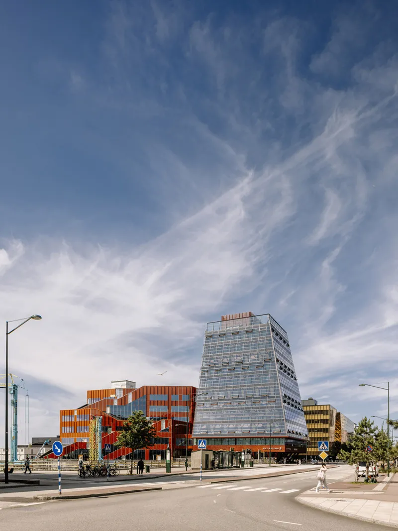 View from the pedestrian boulevard showing the tower and adjacent red perforated volumes under wispy clouds