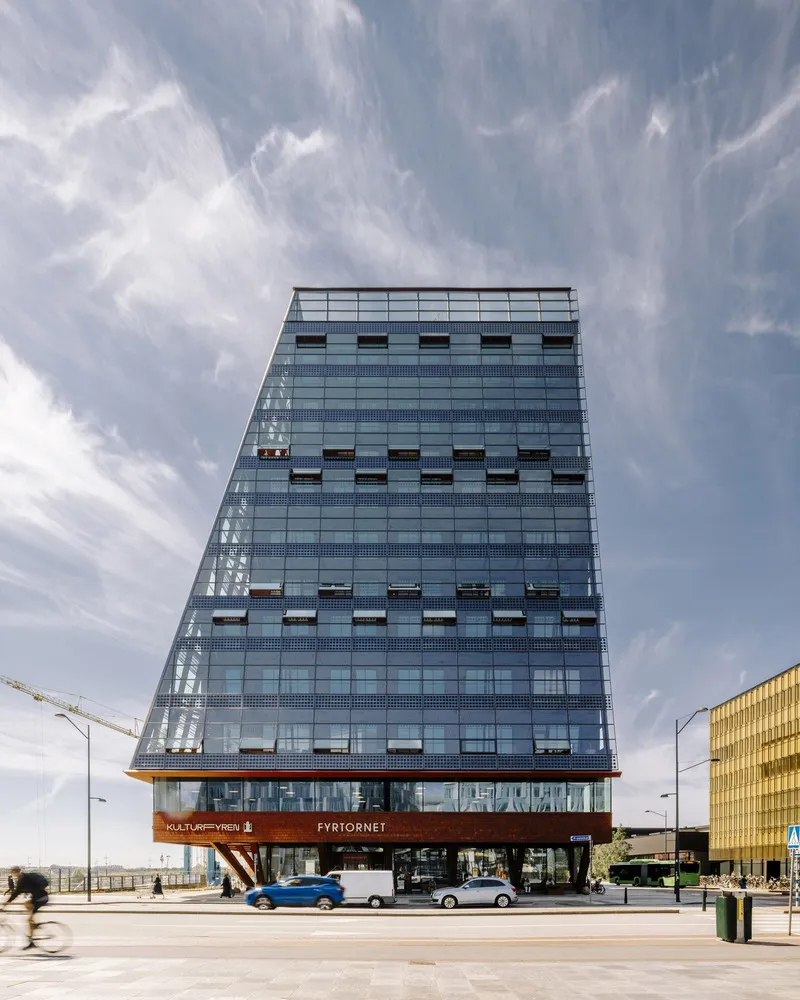 Street-level view of the glass tower facade rising above a double-height red-orange base with vehicles passing below