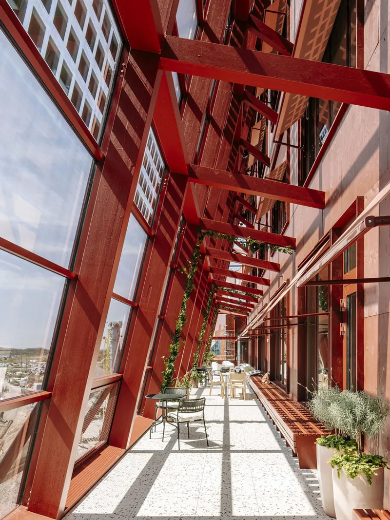 Glazed corridor with angled red steel trusses casting diagonal shadows across the terrazzo floor and planters