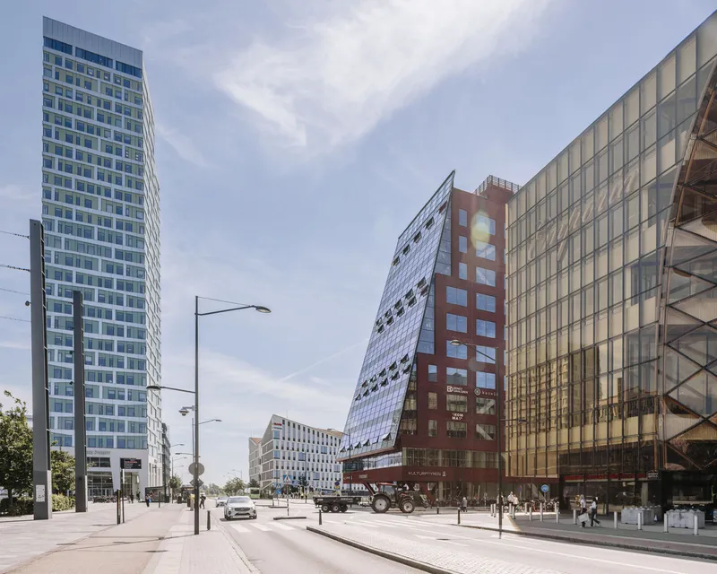 Street view of the mixed-use complex with glass tower and angled metal facade under cloudy skies