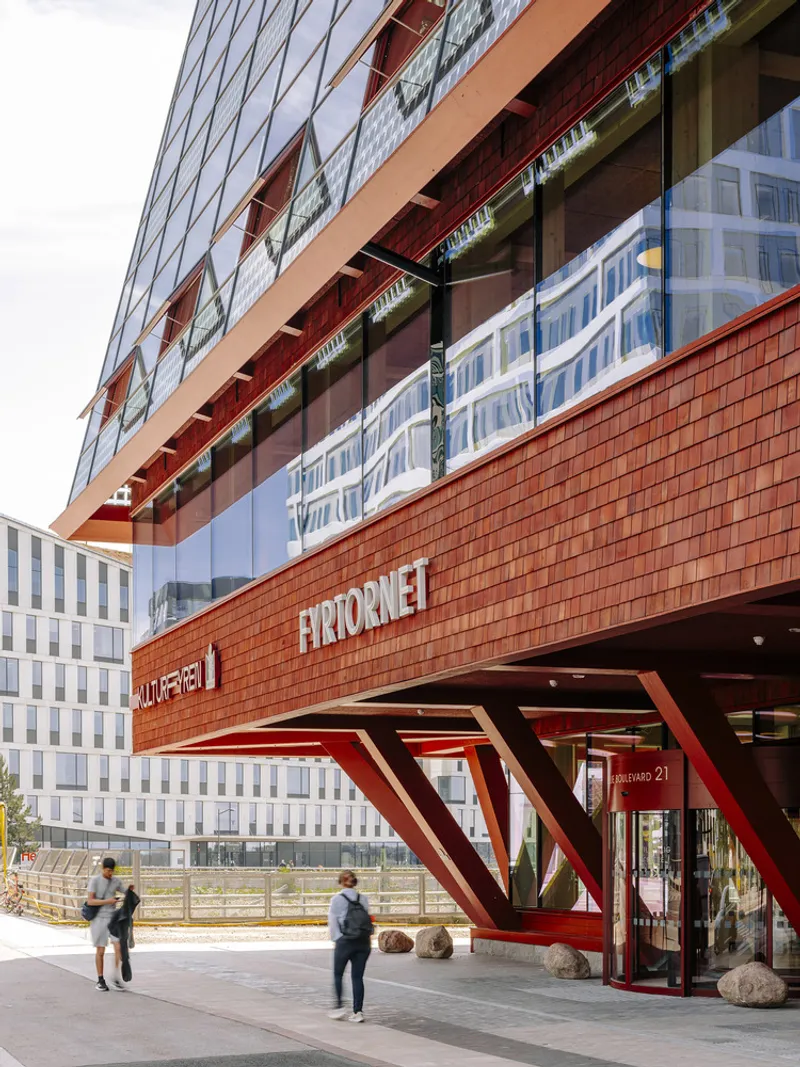 Close-up of the cantilevered podium with red diagonal columns and textured metal cladding above pedestrians