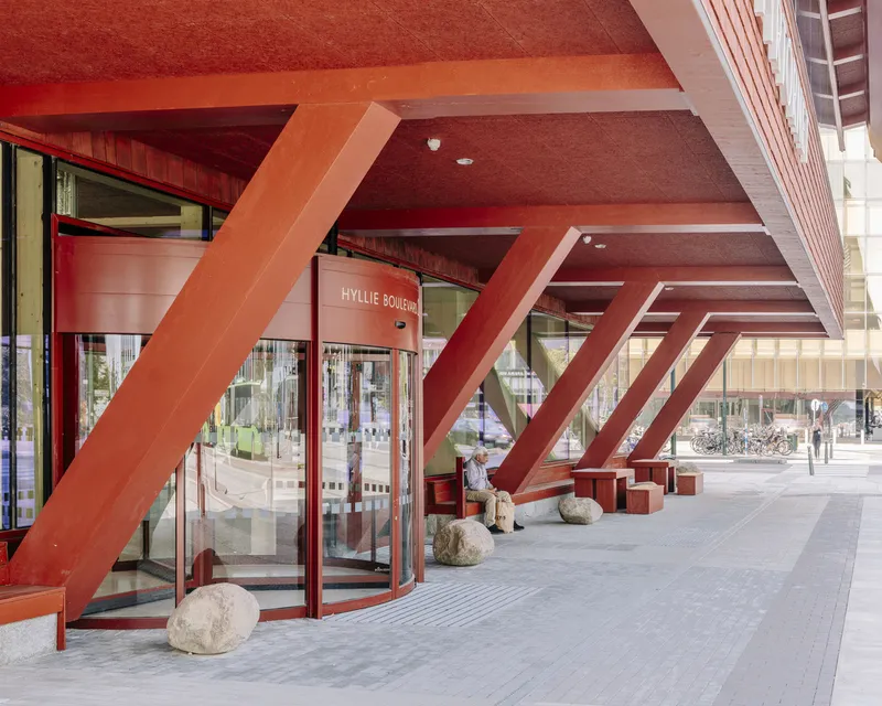 View under the elevated lobby canopy showing red steel braces and glass storefront with person seated
