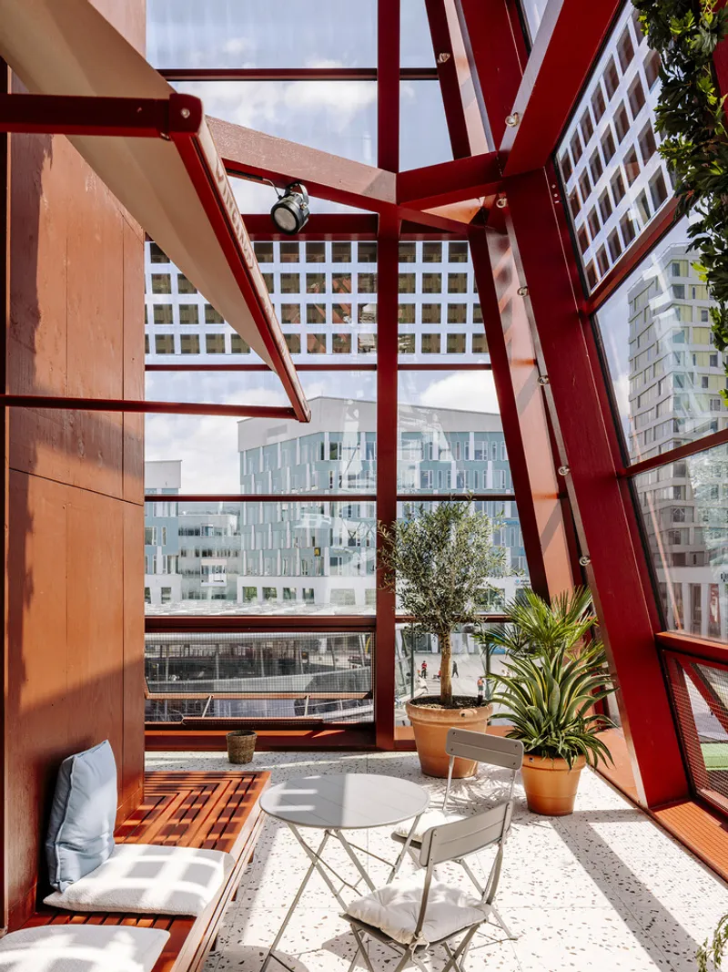Interior balcony with red steel diagonal bracing, glazed facade overlooking the street and potted plants in afternoon sunlight