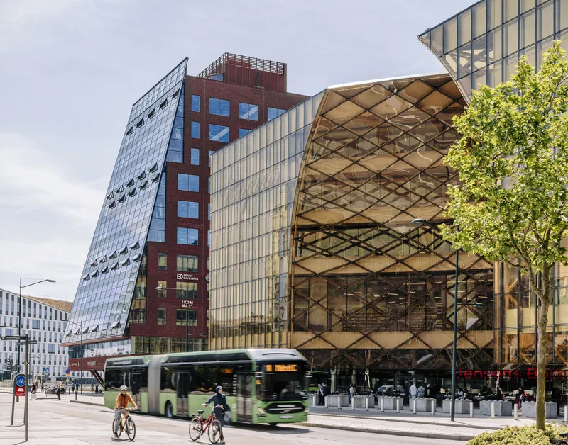 Street view showing faceted glass facade, red brick tower and diamond-patterned timber screen with cyclists and passing bus