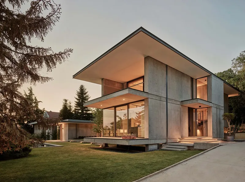 Garden view of the concrete volume with broad roof overhang and full-height glazing at dusk