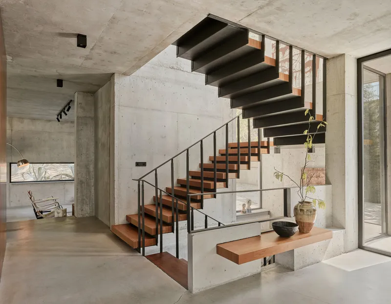 Open-riser timber staircase with steel handrail rising through a double-height concrete-walled entry hall