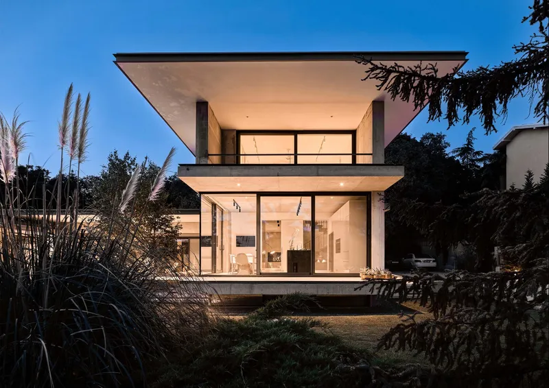 Rear elevation with stacked glazed volumes framed by pampas grass and evergreens at twilight