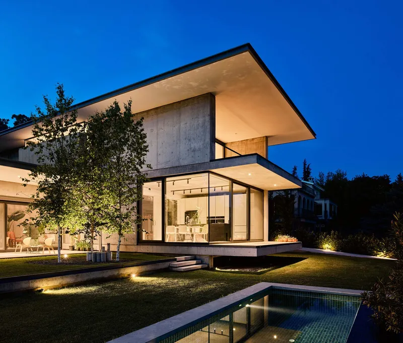 Concrete volume with deep roof overhang and floor-to-ceiling glazing beside the reflecting pool at night